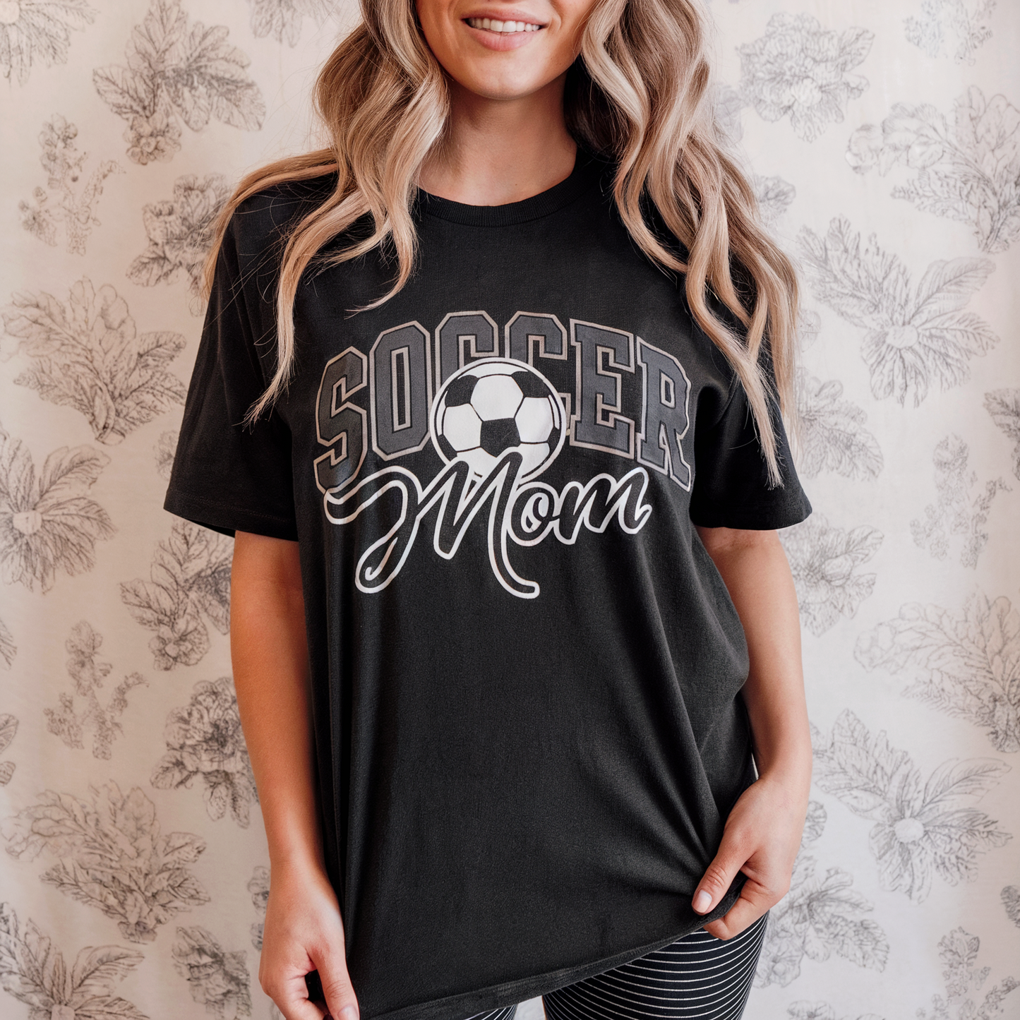 Woman wearing a black 'Soccer Mom' t-shirt against a floral wallpaper background